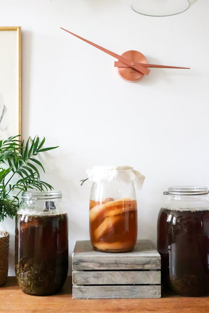 Glass jars with infused liqueurs with ingredients placed on wooden table near green plant near wall with decorations at home