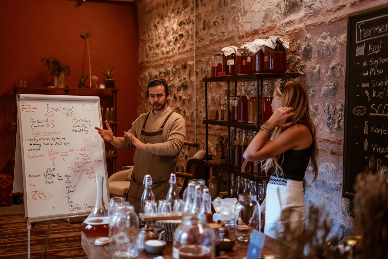 A bearded man in an apron explains fermentation concepts indoors with a whiteboard.